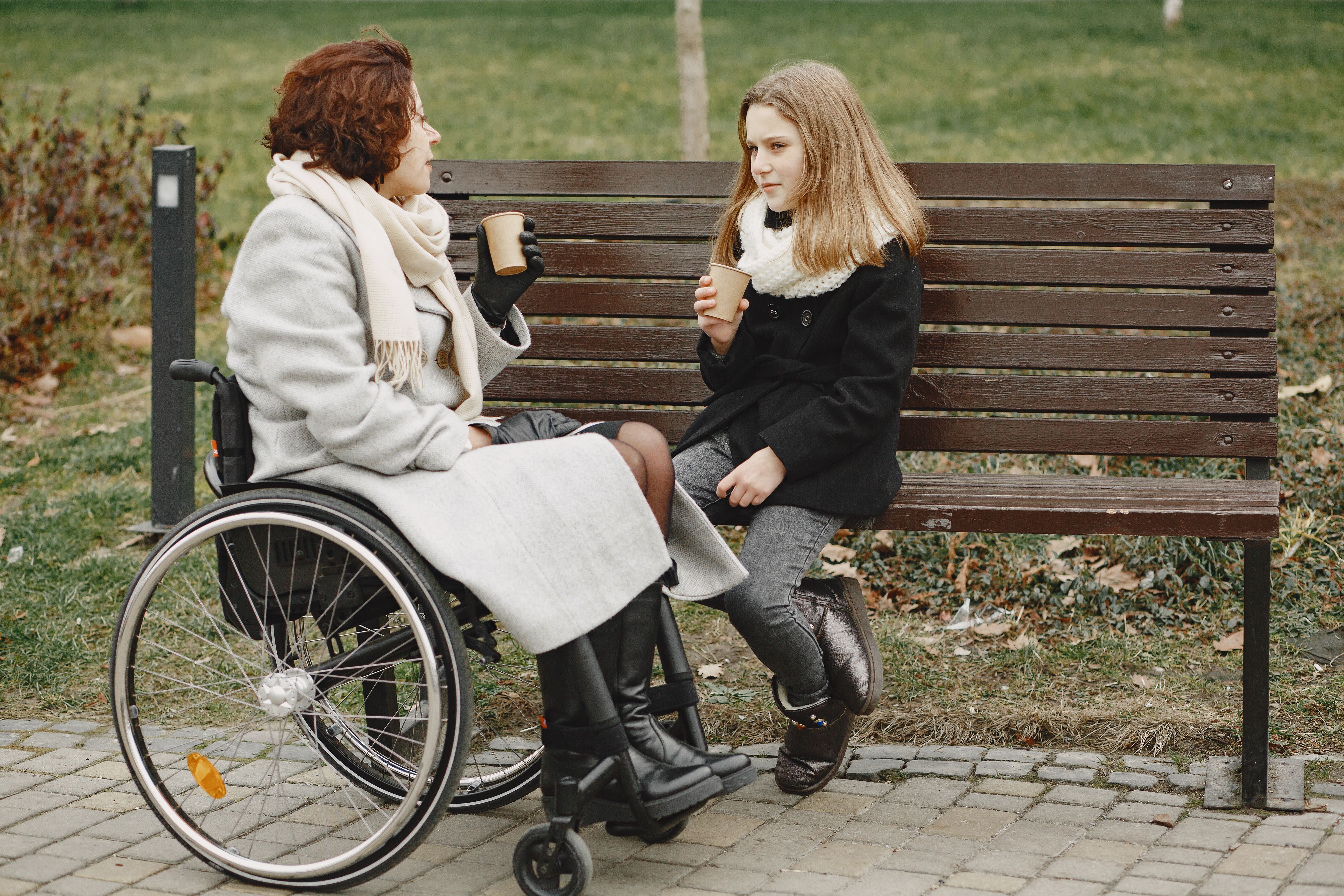 A woman who uses a wheelchair and a younger companion sitting together in a park, talking over warm drinks